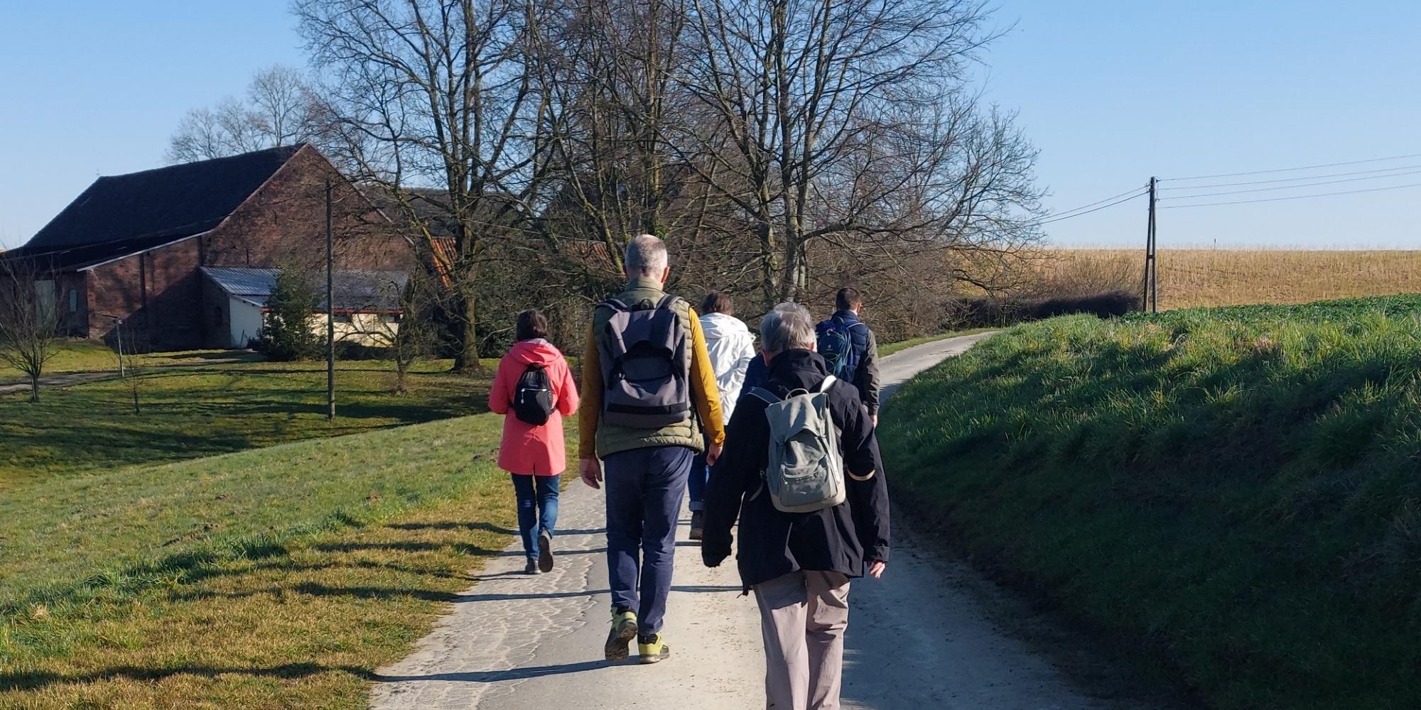 Wanderung Frühjahr 2025 Eine Gruppe von Menschen läuft über einen asphaltierten Feldweg auf ein Gehöft zu. Vor dem Gehöft stehen zwei Bäume, die noch kein Laug tragen.