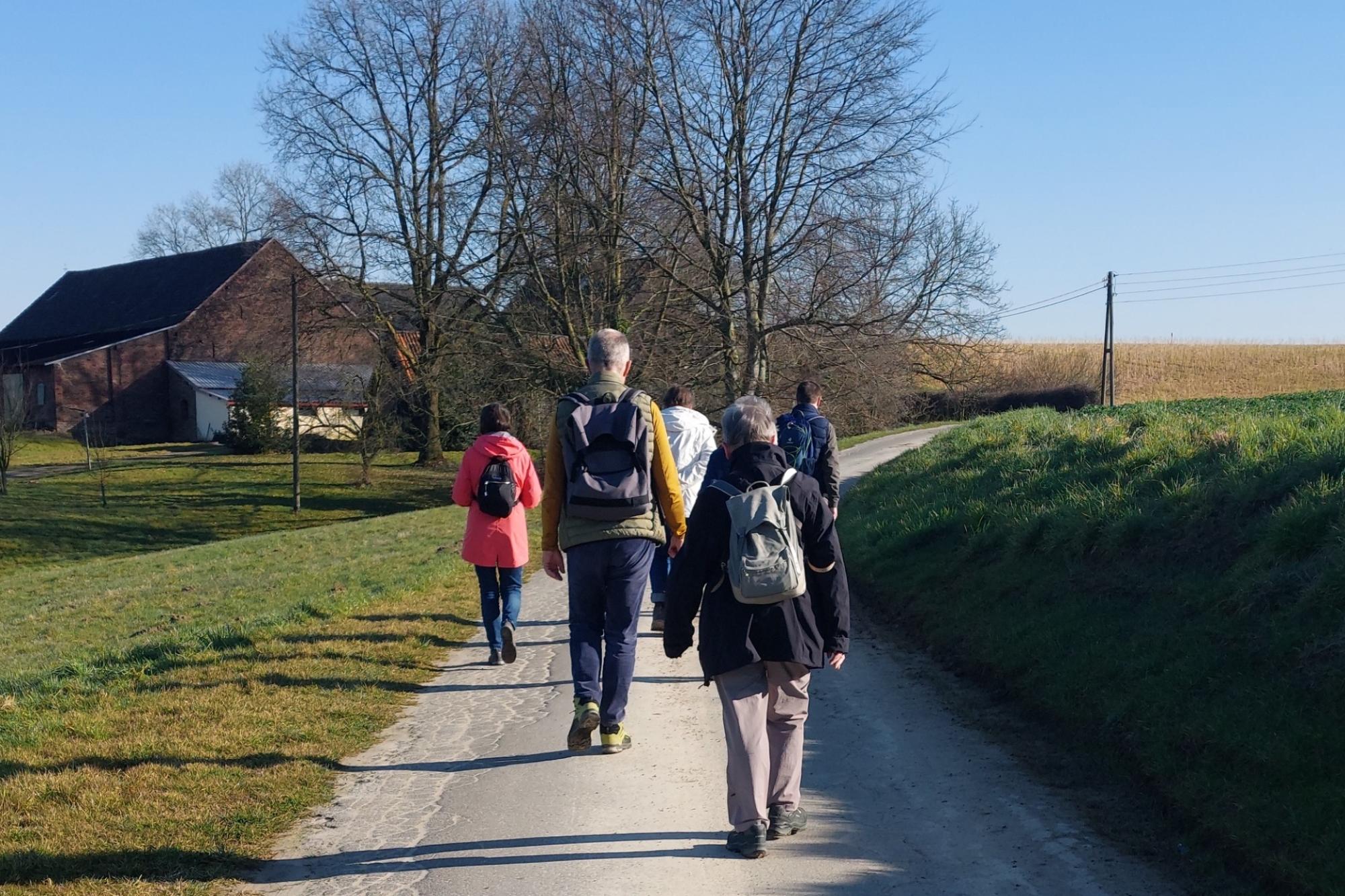 Eine Gruppe von Menschen läuft über einen asphaltierten Feldweg auf ein Gehöft zu. Vor dem Gehöft stehen zwei Bäume, die noch kein Laug tragen.