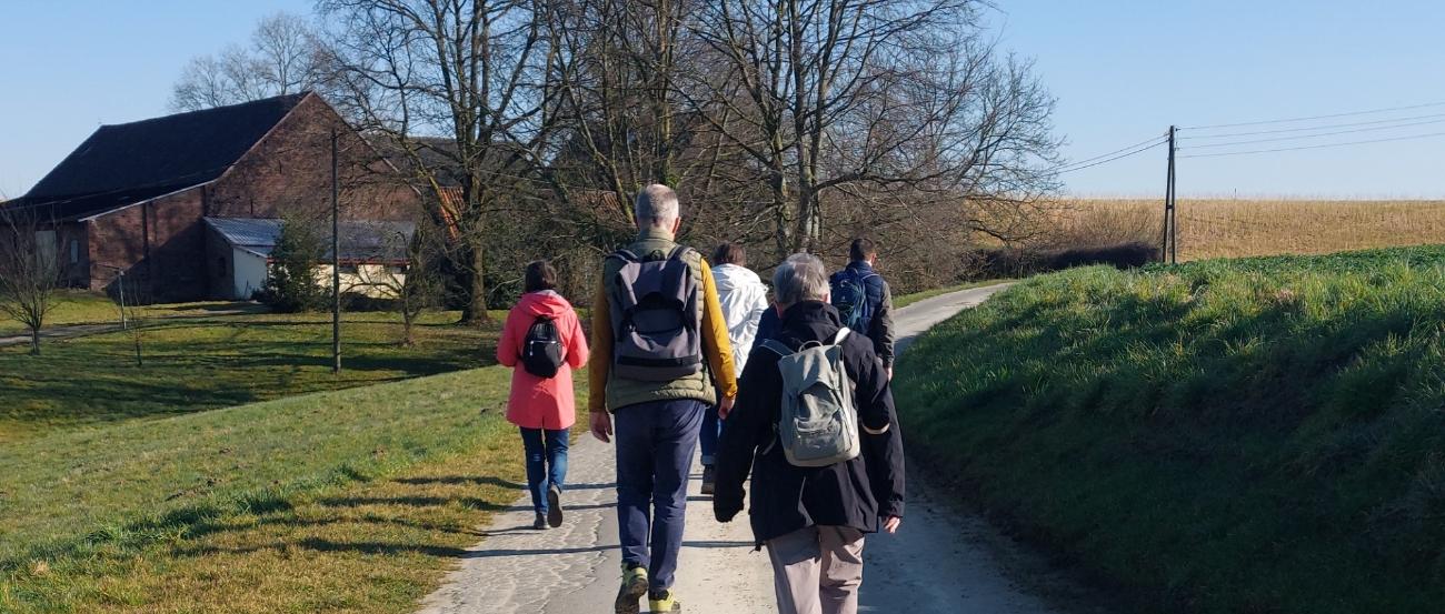 Eine Gruppe von Menschen läuft über einen asphaltierten Feldweg auf ein Gehöft zu. Vor dem Gehöft stehen zwei Bäume, die noch kein Laug tragen.