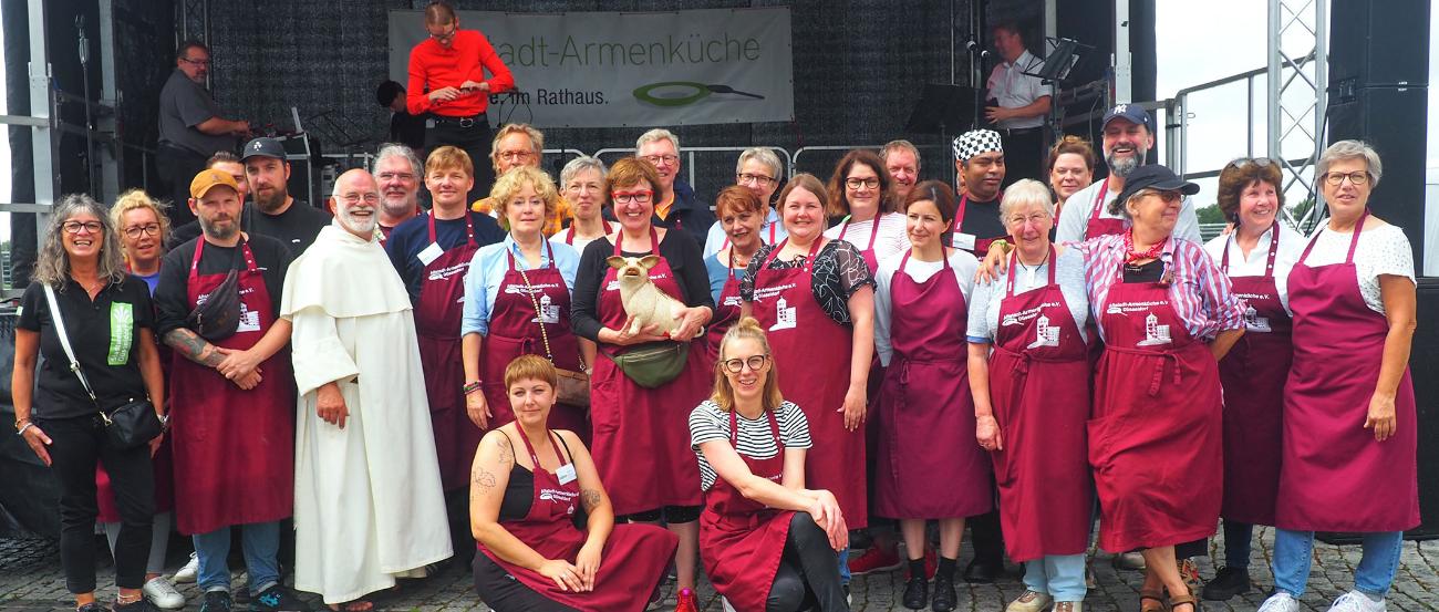 Altstadt Armenküche Ehrenamtliche (c) Altstadt-Armenküche Pater Wolfgang und die Ehrenämtler der Armenküche stehen gemeinsam vor einer Bühne und lächeln.